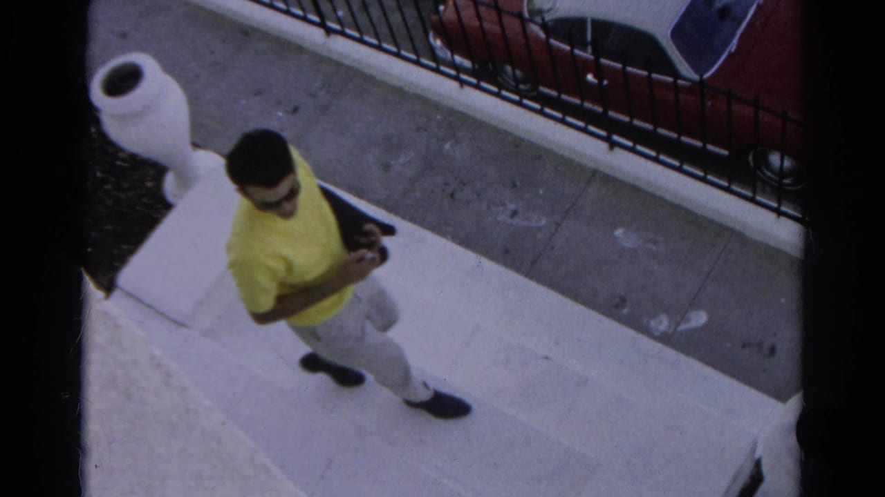 In 1966 Silver Lake, a young man in a yellow shirt and sunglasses descends concrete steps near a sidewalk, iron fence, and red car.