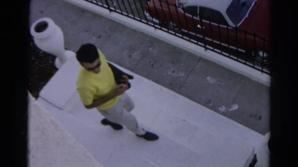 In 1966 Silver Lake, a young man in a yellow shirt and sunglasses descends concrete steps near a sidewalk, iron fence, and red car.