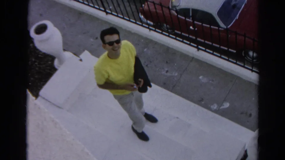 In Silver Lake home-movie footage, the young man looks up and smiles from the steps, holding a dark jacket near a sidewalk and fence.