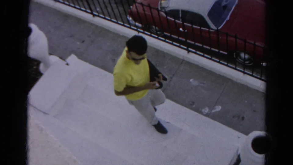 The 1966 home-movie frame shows the man mid-step on concrete stairs, with the sidewalk, black fence, and red parked car behind him.