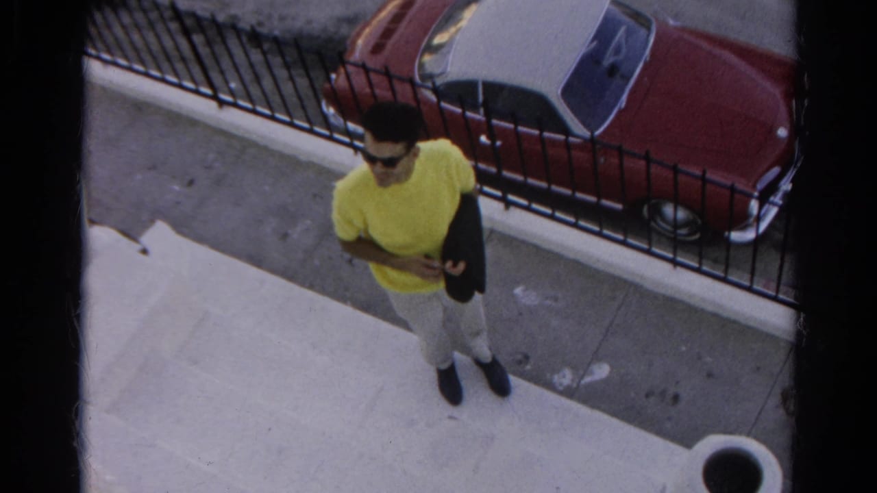 Man in a yellow T-shirt and sunglasses stands on a sidewalk near a red-and-white convertible behind a fence.