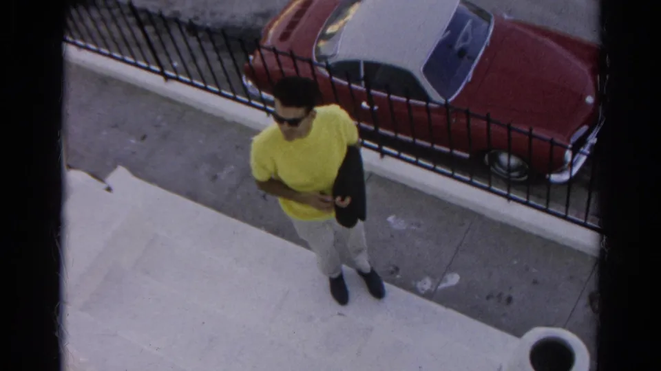 In 1966 Silver Lake footage, a man in a yellow T-shirt holds a dark jacket on a sidewalk beside a black fence and parked convertible.