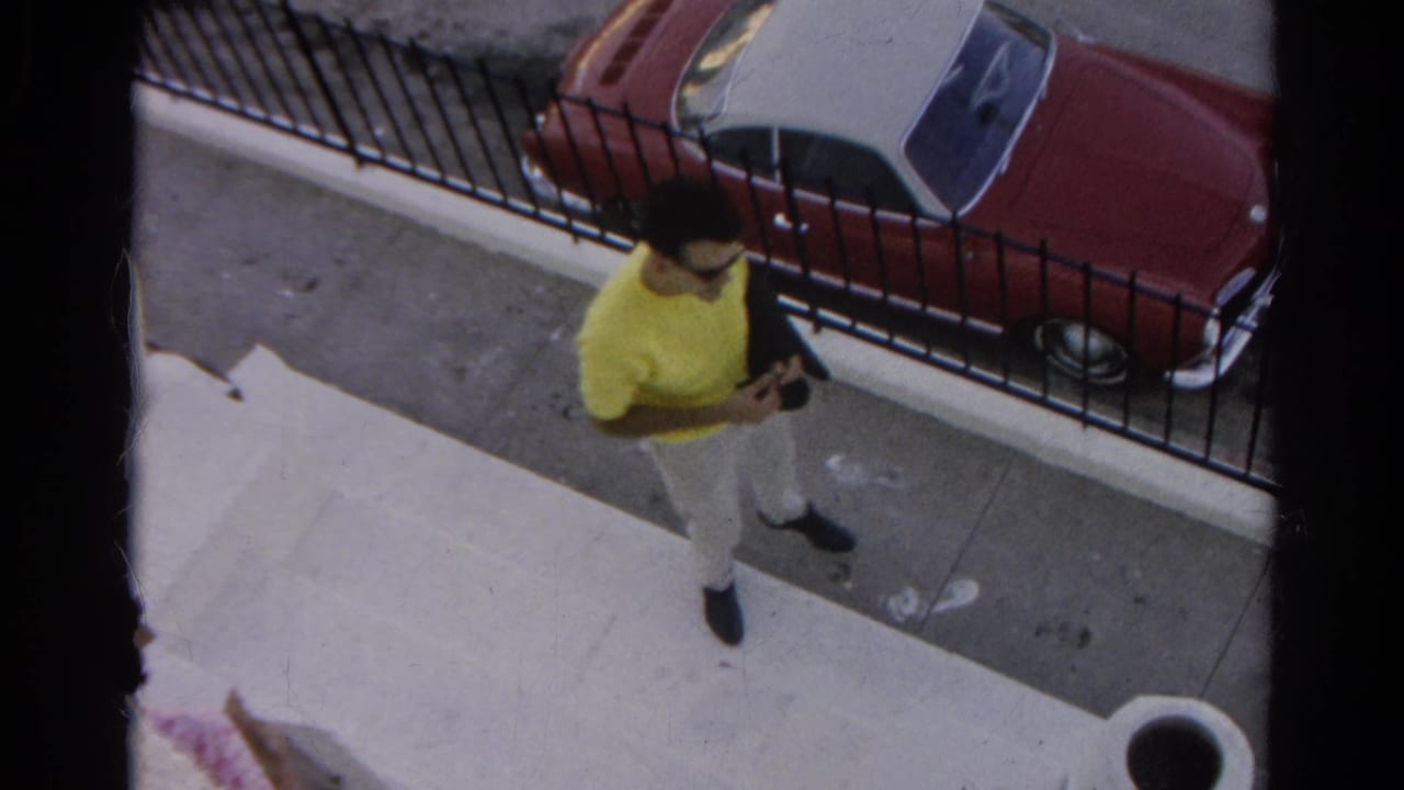 Man in a yellow T-shirt walks along the sidewalk while holding a dark jacket near the parked convertible.