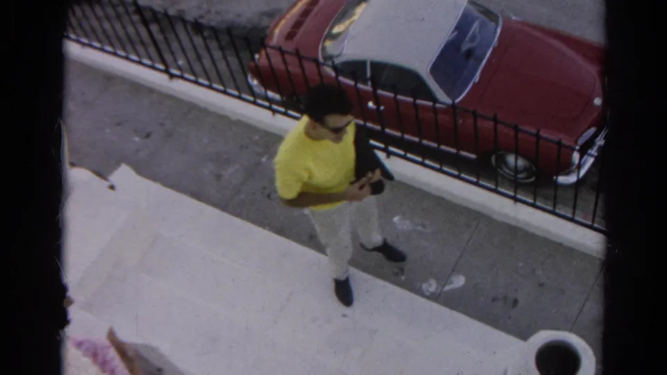 The man in yellow moves along a Silver Lake sidewalk, holding a dark jacket with the red-and-white convertible still visible behind the fence.