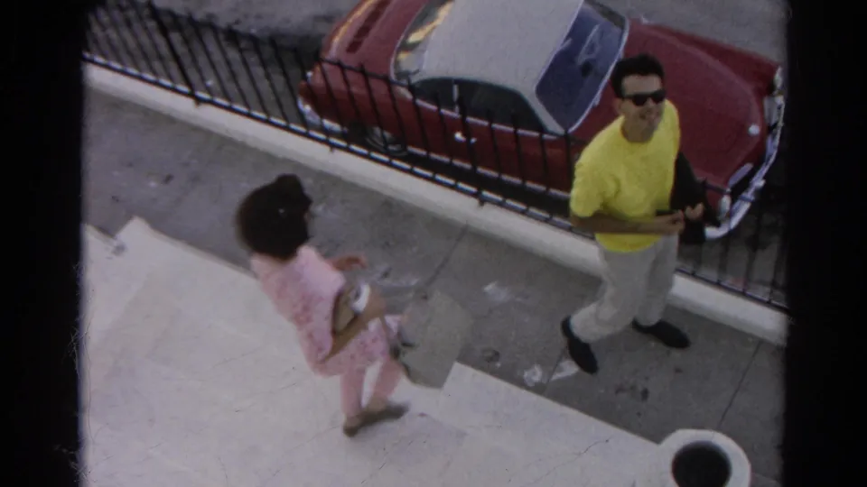 A woman in a pink patterned outfit carrying a purse passes the man in yellow on a Silver Lake sidewalk near the parked convertible.