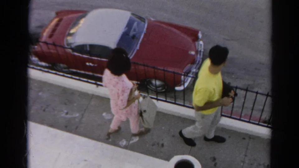 The man in yellow and woman in pink separate along the Silver Lake sidewalk, with the black fence and red-and-white convertible behind them.