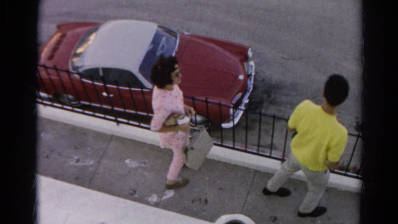 Man and woman walk beside a fence near a red and white parked car.