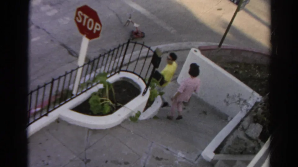 The couple walks away along the Silver Lake sidewalk, with the stop sign, planter, black fence, and low wall still visible.