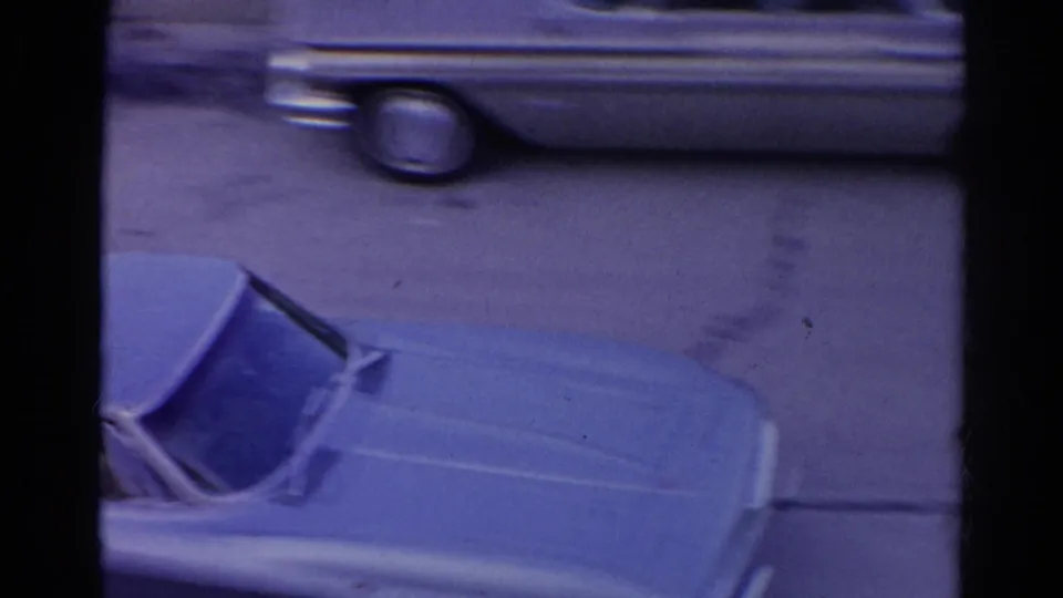 Close 8mm view of the blue convertible on a Silver Lake street, partly obscured by window framing and a passing pale car.