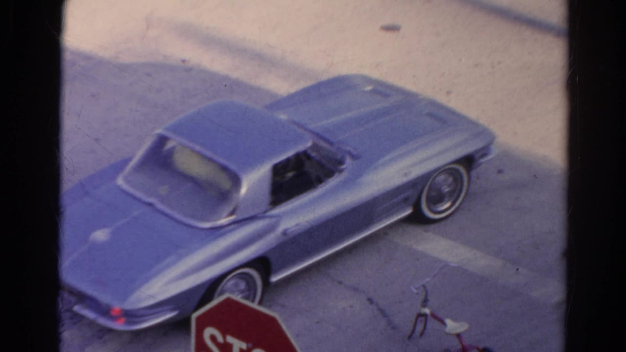 The blue classic sports car turns through a Silver Lake intersection, with a stop sign edge, curb, shadows, and 8mm film grain visible.