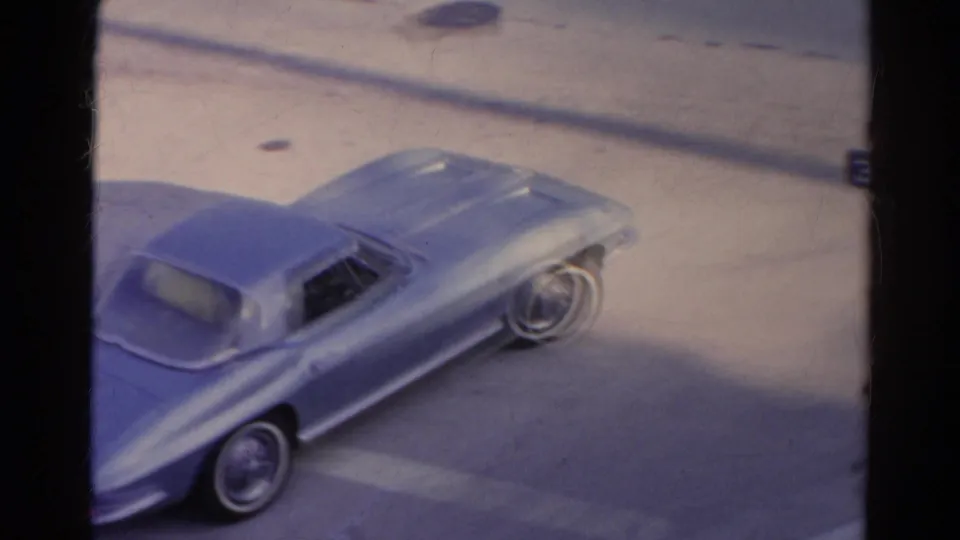 The light blue sports car continues its turn in the 1966 Silver Lake street scene, viewed from above with soft 8mm grain and muted color.