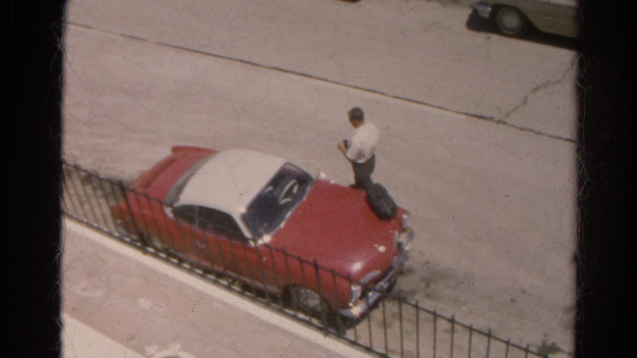 Man in a white shirt stands near a red and white car parked by a black metal fence.