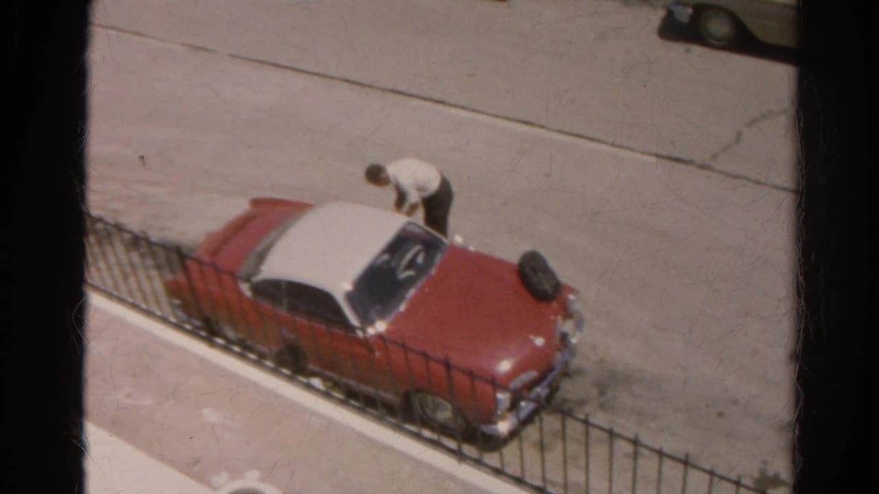 Man bends beside the red and white car, with the street and another parked car behind him.