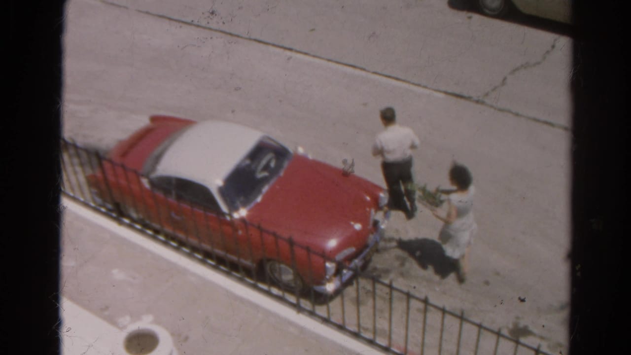 Two adults stand beside a red car with a white roof on a sunlit street.