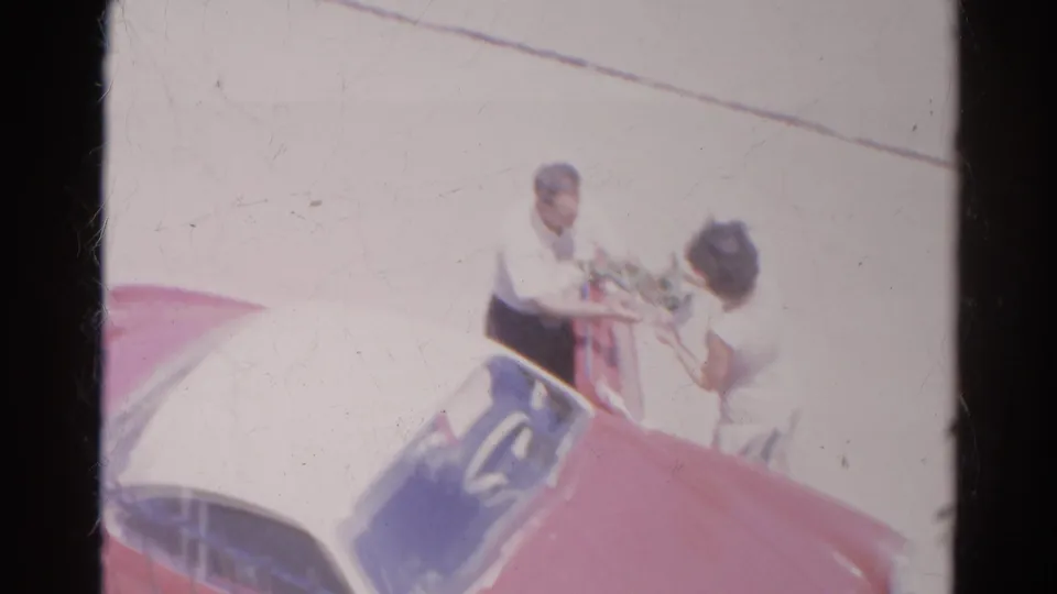 The woman appears to offer an item to the man beside the front of the red car in the Silver Lake home-movie street scene.