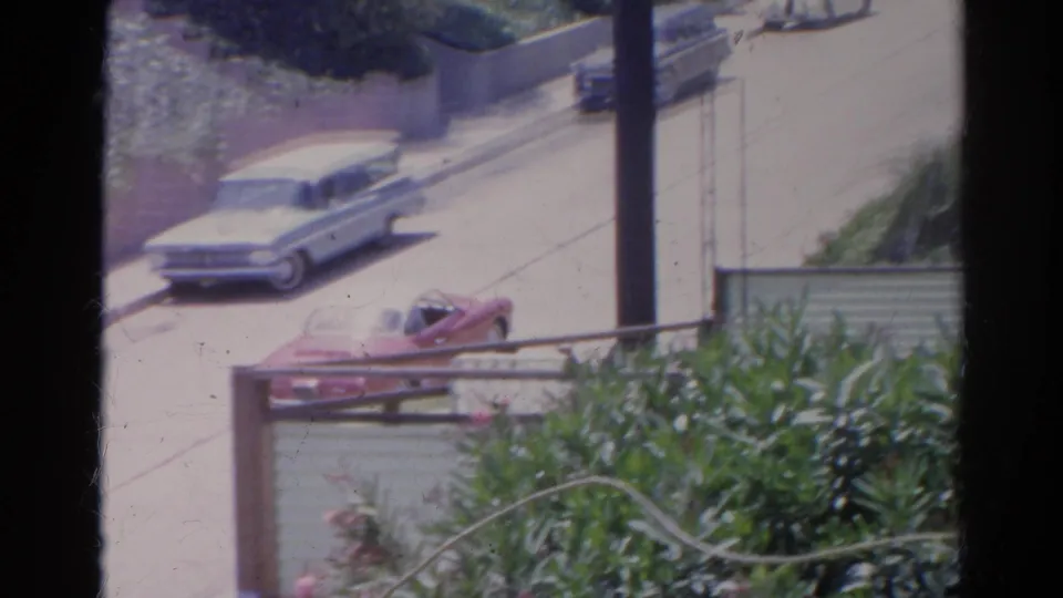 Red convertible driving below an elevated viewpoint near a pale station wagon on a 1966 Silver Lake residential street.