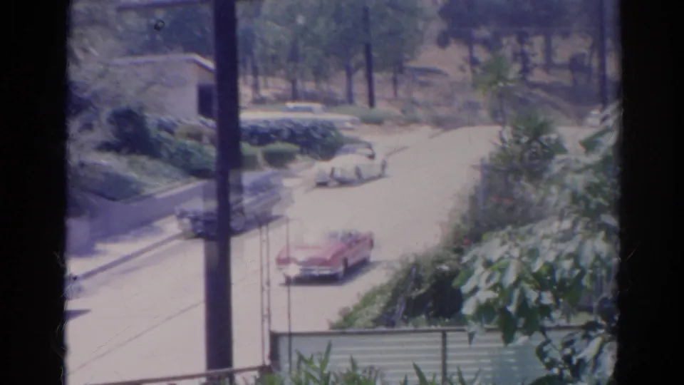 Elevated 1966 Silver Lake street view with a red convertible, pale station wagon, utility pole, and dense roadside plants.