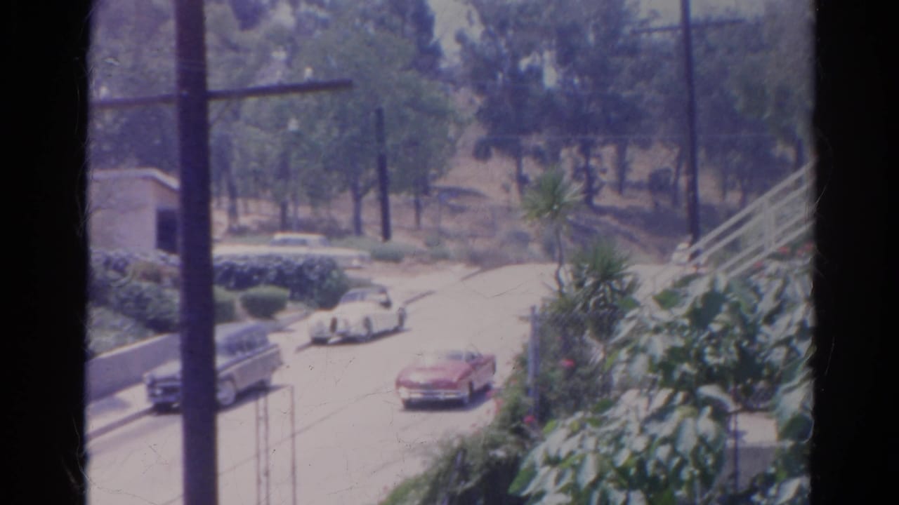 Elevated view of a sunny residential street with a red car, parked cars, trees, and utility poles.