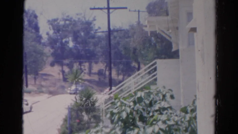 1966 Silver Lake home-movie frame with a white car on a residential road beside trees, utility poles, shrubs, and a white building edge.