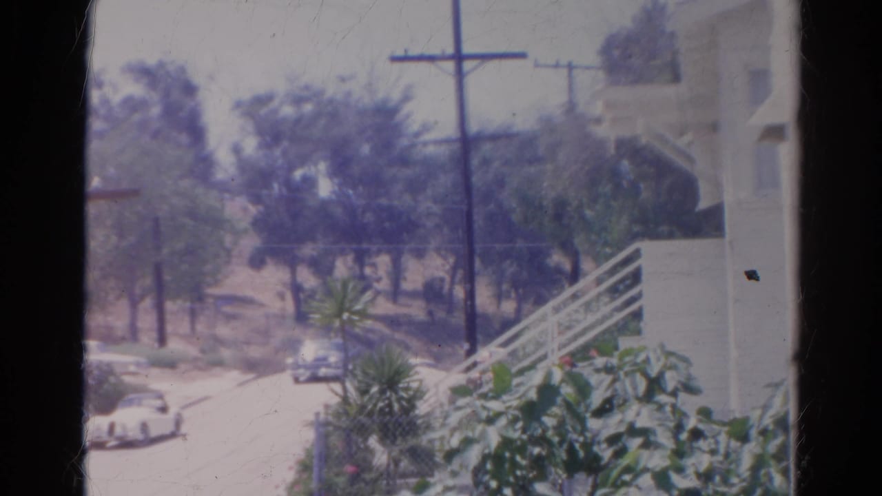 1966 Silver Lake home-movie frame showing cars on a quiet residential street with trees, utility poles, and hillside buildings.