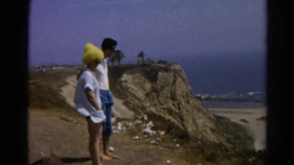 1966 California home-movie frame of two adults at a coastal bluff, with beach, ocean, and distant palm trees visible.