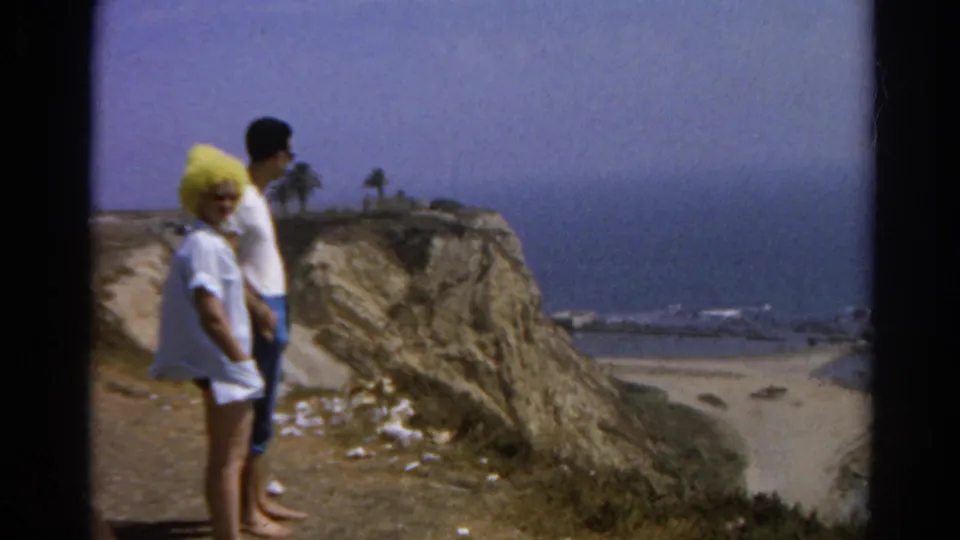 1966 California beach overlook scene with two adults beside a bluff, ocean water, sand, and distant shore structures.