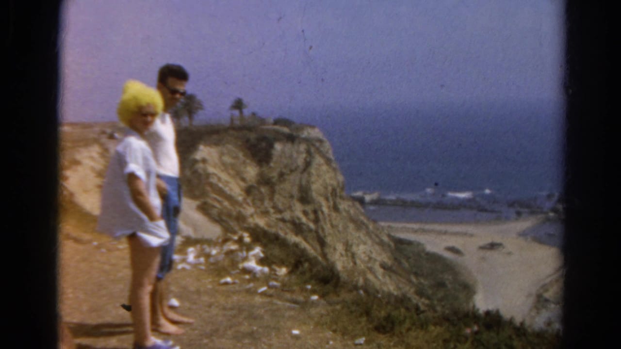 1966 8mm frame of a couple at a California beach overlook, with surf below and the woman in a bright yellow puffy hat.