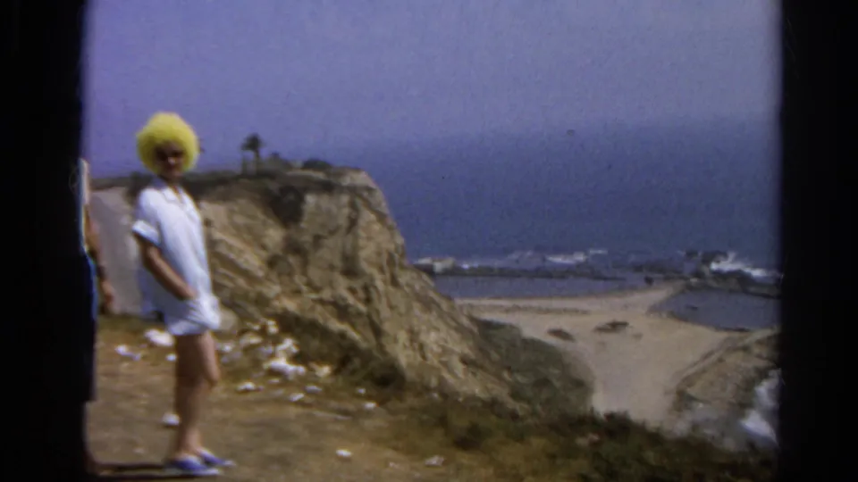 1966 8mm frame of the woman in a yellow hat and white dress standing at a California beach overlook with surf below.