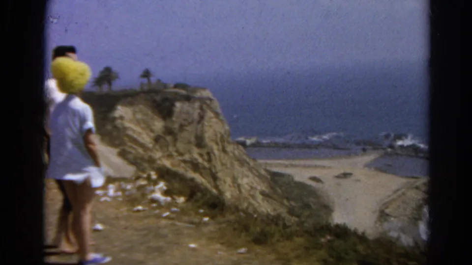 1966 8mm frame of the couple facing a California coastal view from a cliff, with waves, palms, and beach below.
