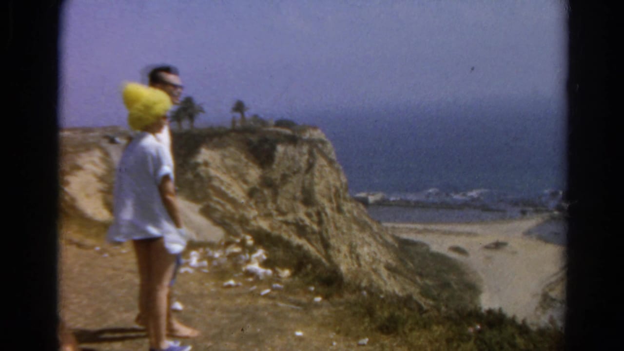 Couple standing on a cliff path above the ocean, with the woman wearing a bright yellow hat.