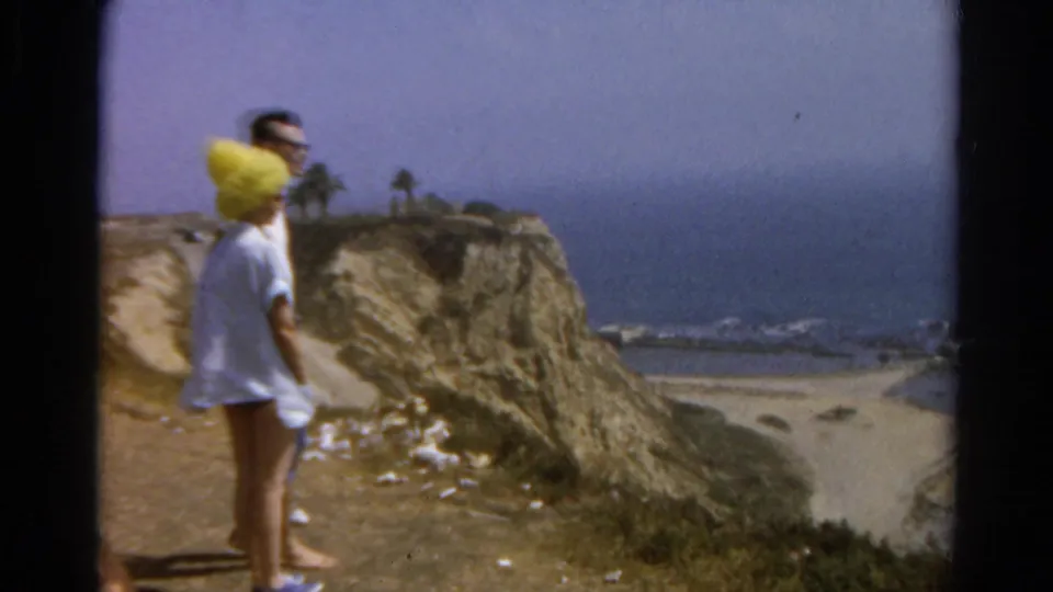 1966 8mm frame of an adult couple on a California coastal cliff path, with ocean, beach, and palm trees in the distance.