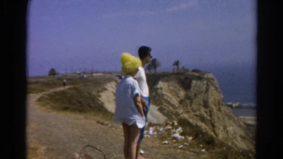 The couple stands on a dirt overlook path in 1966 California home-movie footage, facing the ocean and eroded cliff edge.
