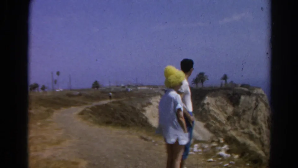 A 1966 8mm frame shows the couple moving along a coastal path, with the cliff face, ocean horizon, and palms beyond.