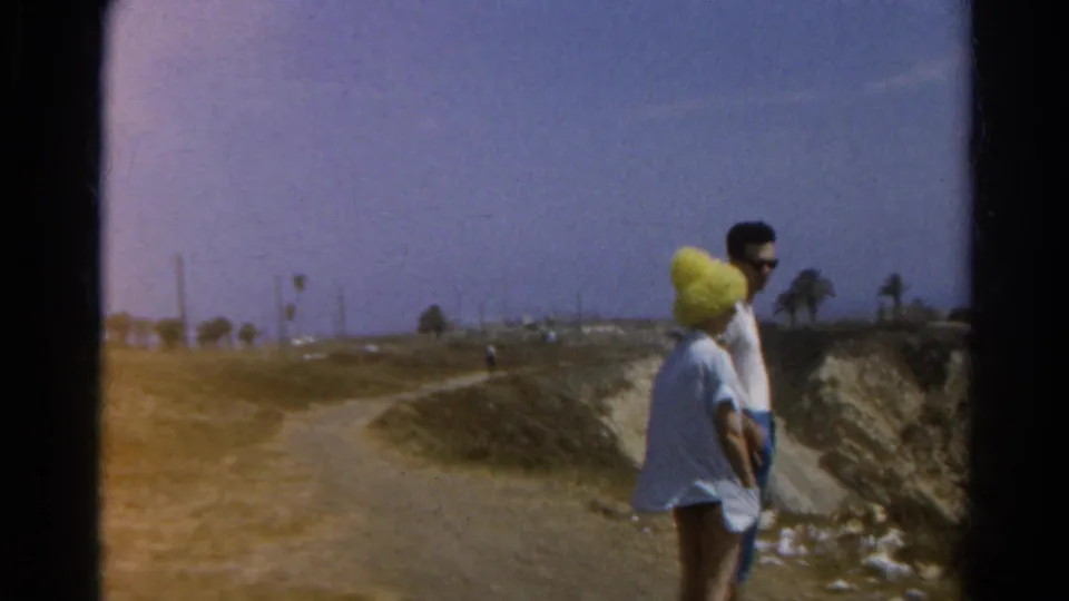 The couple remains beside the coastal path in 1966 California footage, with beach, palms, and eroded bluff visible behind them.