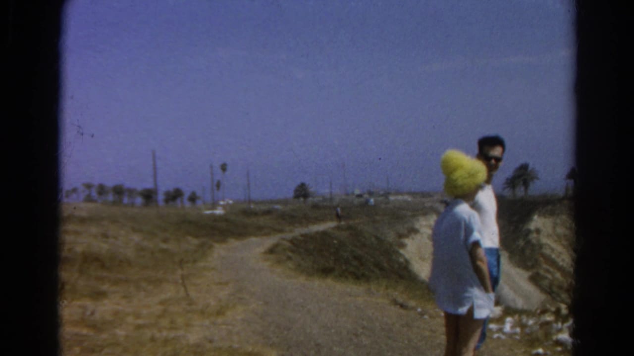 Two adults stand on a dirt overlook trail near eroded cliffs and distant palms.