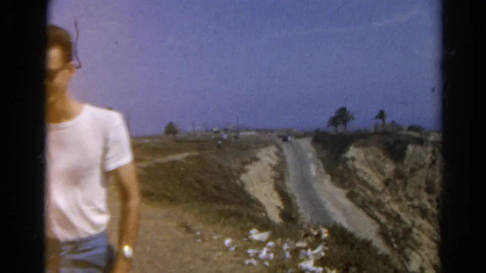 A man in sunglasses and a white T-shirt is partly cropped in a 1966 8mm frame, with a dry cliff road in the background.