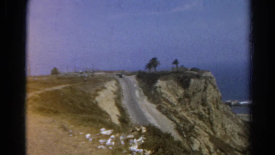 The final 1966 8mm frame holds on the dry cliffside road, palms, and distant water after the adults leave the view.