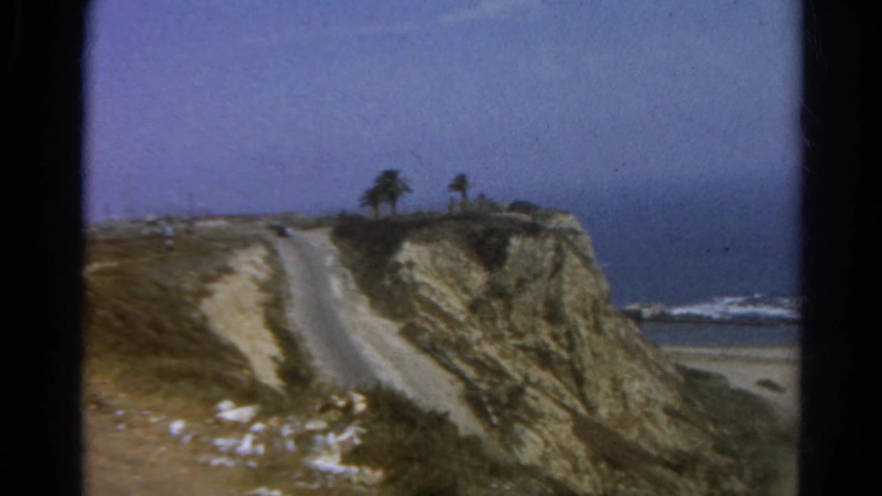 Cliffside road above a sandy beach with ocean waves and small palms near the bluff edge.