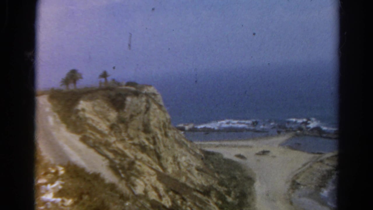 Rocky cliff and pale road overlooking a beach, ocean, and low waves under hazy sky.