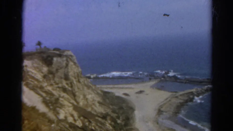 1966 8mm home-movie frame of a California beach overlook, with the camera panning from cliff face toward surf.