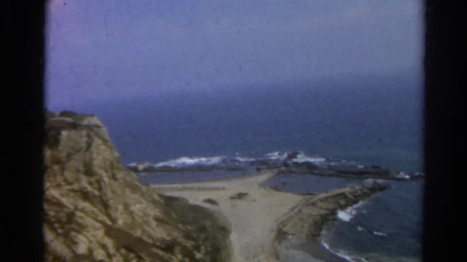 1966 8mm coastal landscape labeled California, showing beach, surf, ocean, and cliff edge during a slow pan.