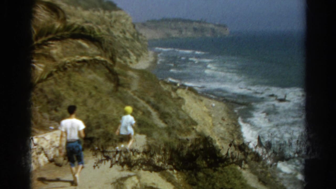 Two people walk down a dirt path beside rocky ocean cliffs.