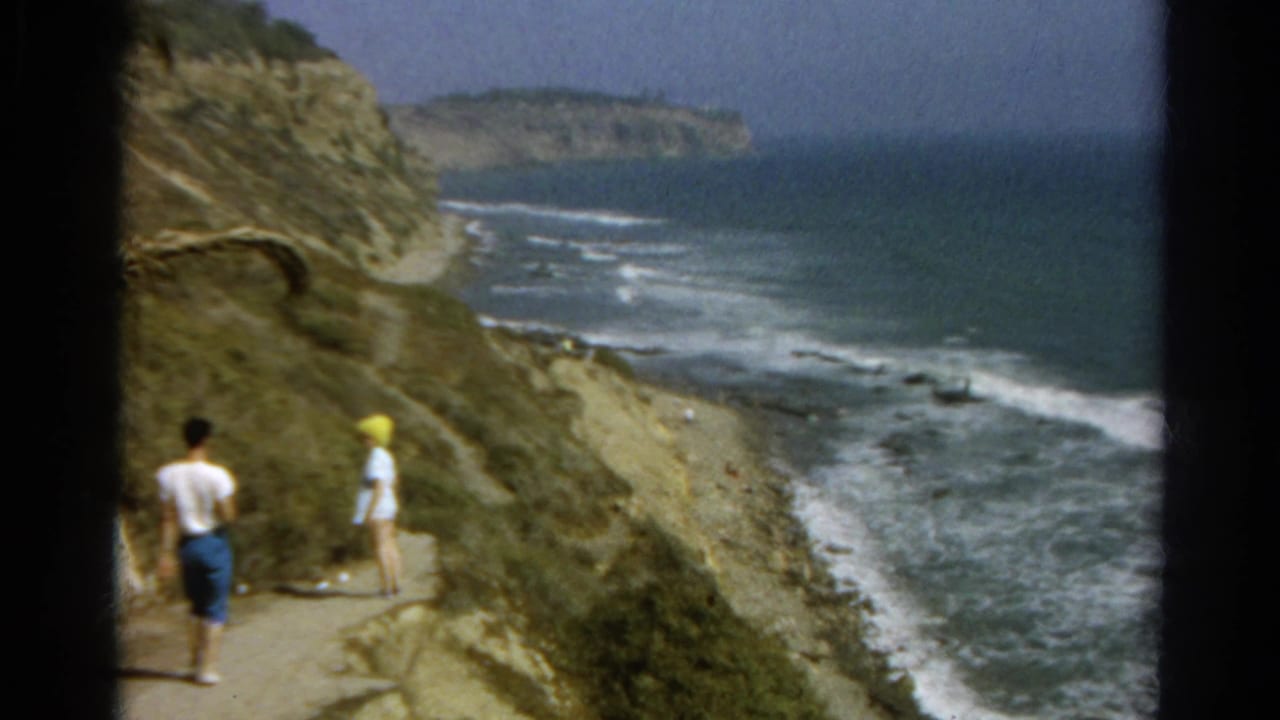 A white-shirted walker and a person in a yellow hat continue along a seaside cliff trail in 1960s 8mm footage.