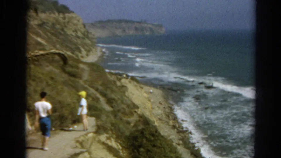 A white-shirted walker and a person in a yellow hat continue along a seaside cliff trail in 1960s 8mm footage.