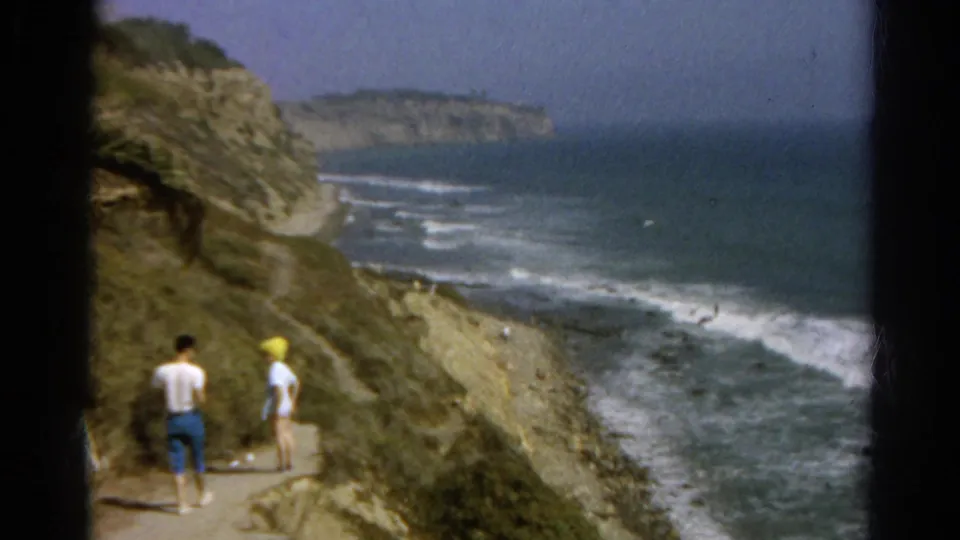 The walkers are seen from behind on a dirt path with steep rocks and rolling surf below.