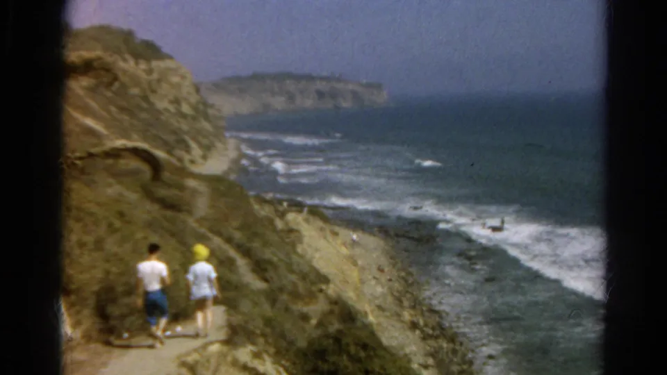 Two figures walk near a bend in a rugged oceanfront trail, with waves and a distant headland visible.