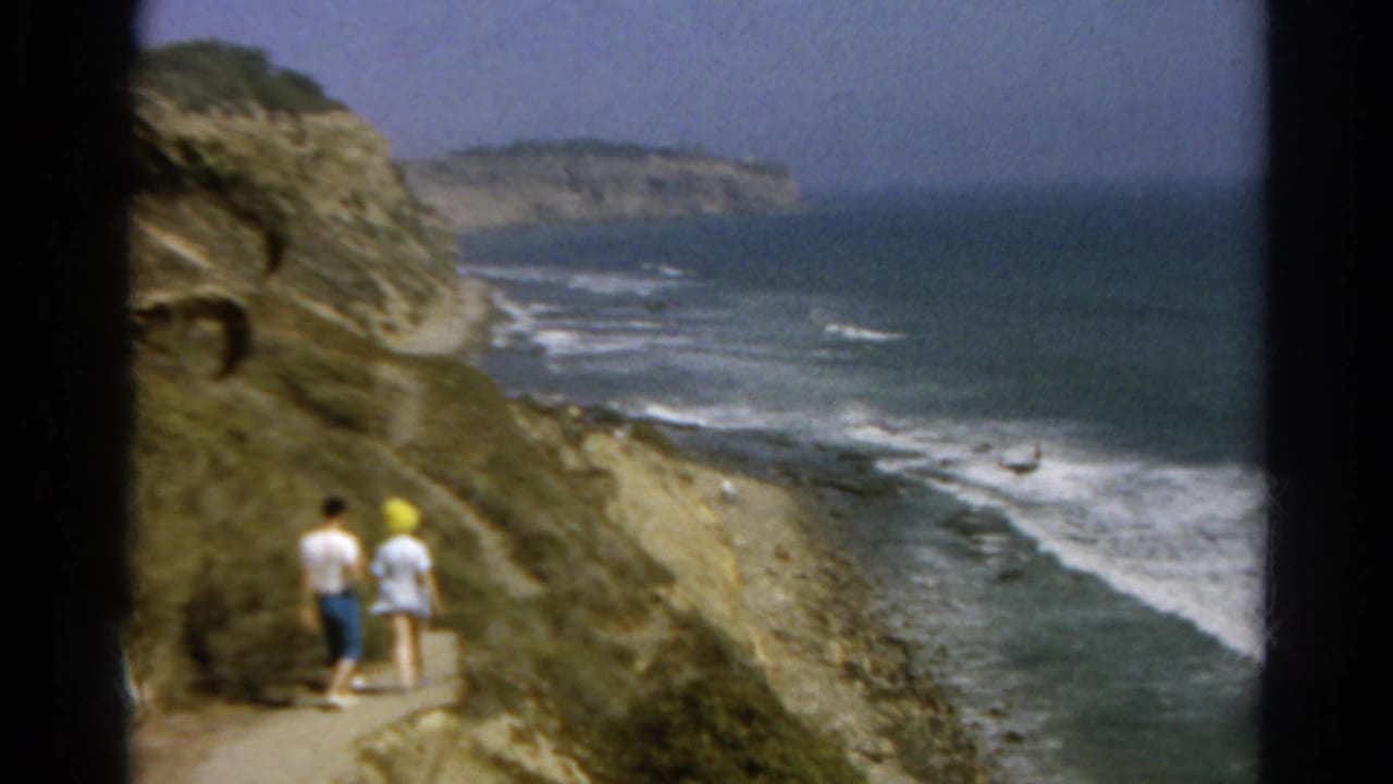 Two adults walk on a dirt cliff path above ocean surf.