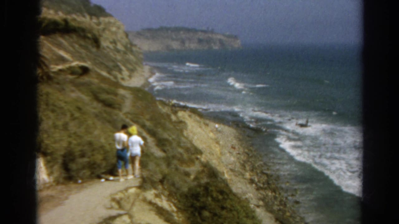 The couple remains on the coastal path, with rocky bluff walls, breaking waves, and a distant coastline visible in the 1966 home-movie frame.