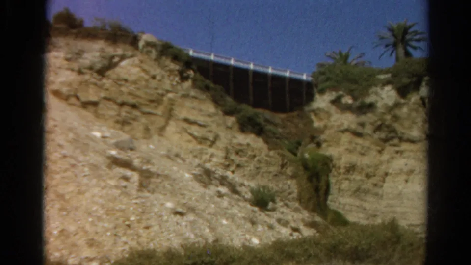 The camera frames the bluff itself, showing eroded rock, a dusty path, sparse vegetation, palm trees, and a fence or bridge-like structure above.