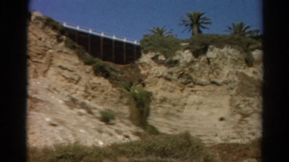 A later frame shows the same coastal bluff area with layered rock, dry vegetation, palms, and a raised fence or bridge-like edge against the sky.